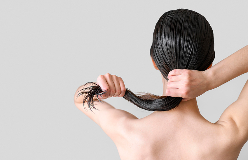 Mujer de espaldas sostiene su cabello h&uacute;medo con ambas manos formando una coleta